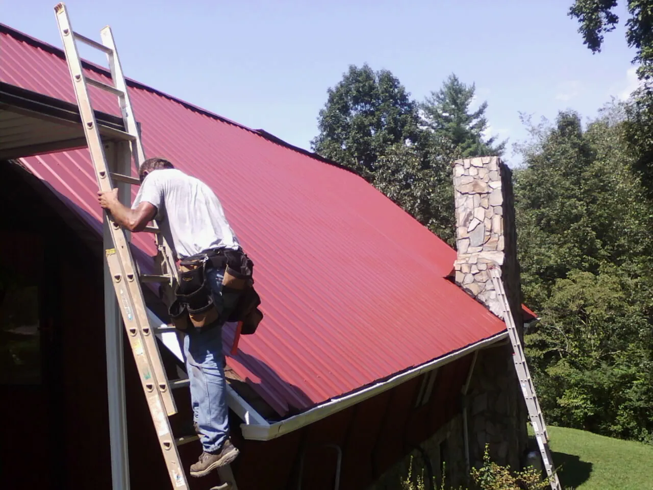 Roofer on a ladder beside a red metal roof and stone chimney — Soffit & Fascia Repair by Roofing Brothers in Big Piney